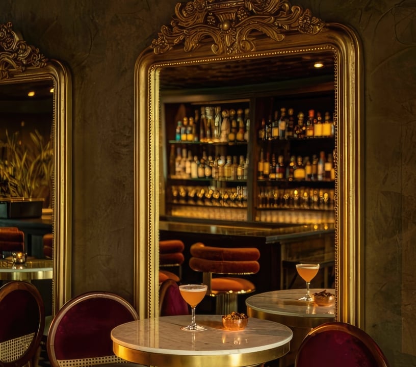 A small table with elegant chairs at Darling. A cocktail is placed on the table and a view of the bar is seen through a mirror behind the setting.