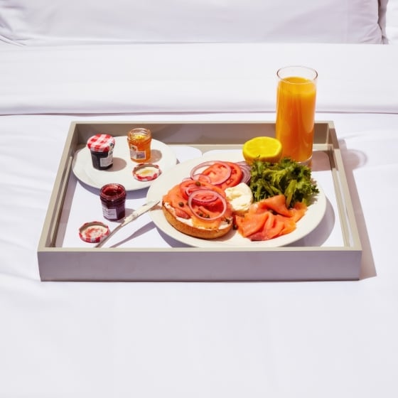 An in-room dining breakfast tray of a smoked salmon bagel, jams, and orange juice on a bed.