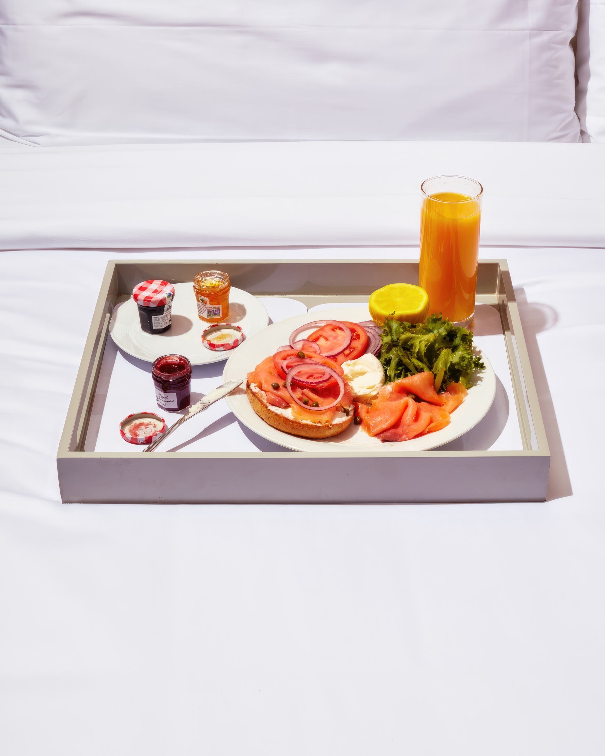 An in-room dining breakfast tray of a smoked salmon bagel, jams, and orange juice on a bed.