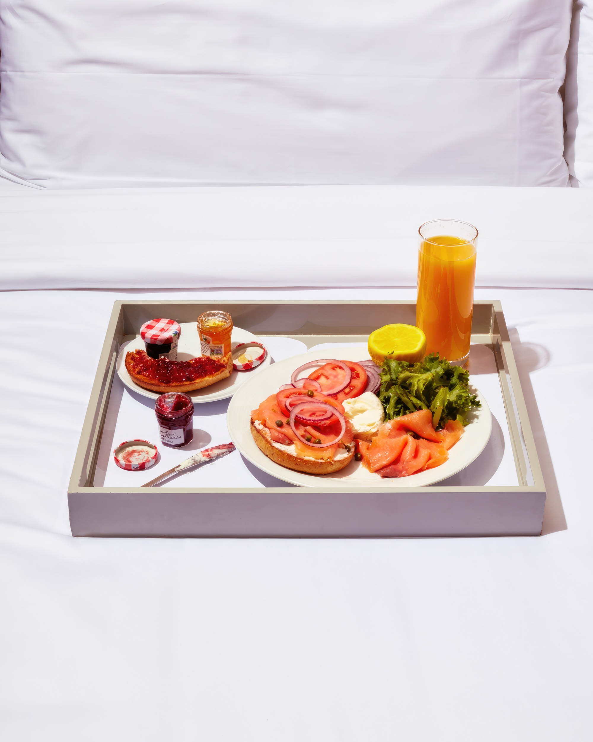 An in-room dining breakfast tray of a smoked salmon bagel, jams, and orange juice on a bed.