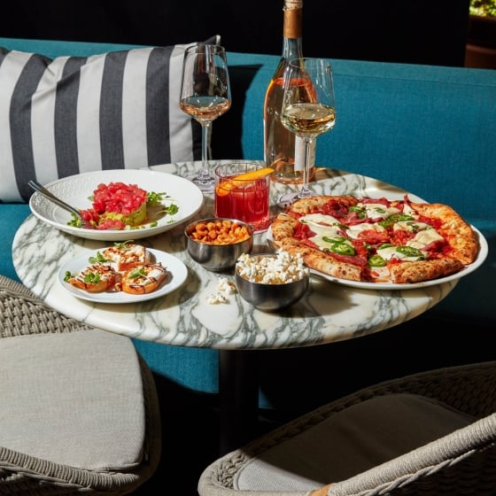 A restaurant table holds pizza, appetizers, tuna tartare, wine and cocktails.