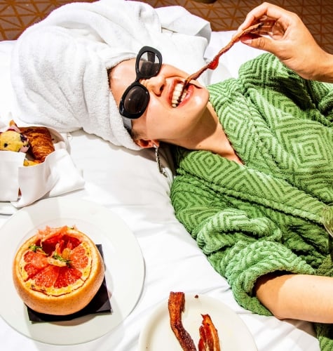 A woman in a green robe and sunglasses eats a piece of bacon while laying down in bed.