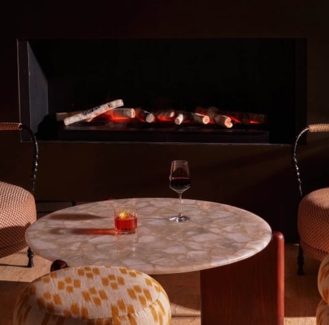 A glass of wine and a cocktail on a table in front of a fireplace.